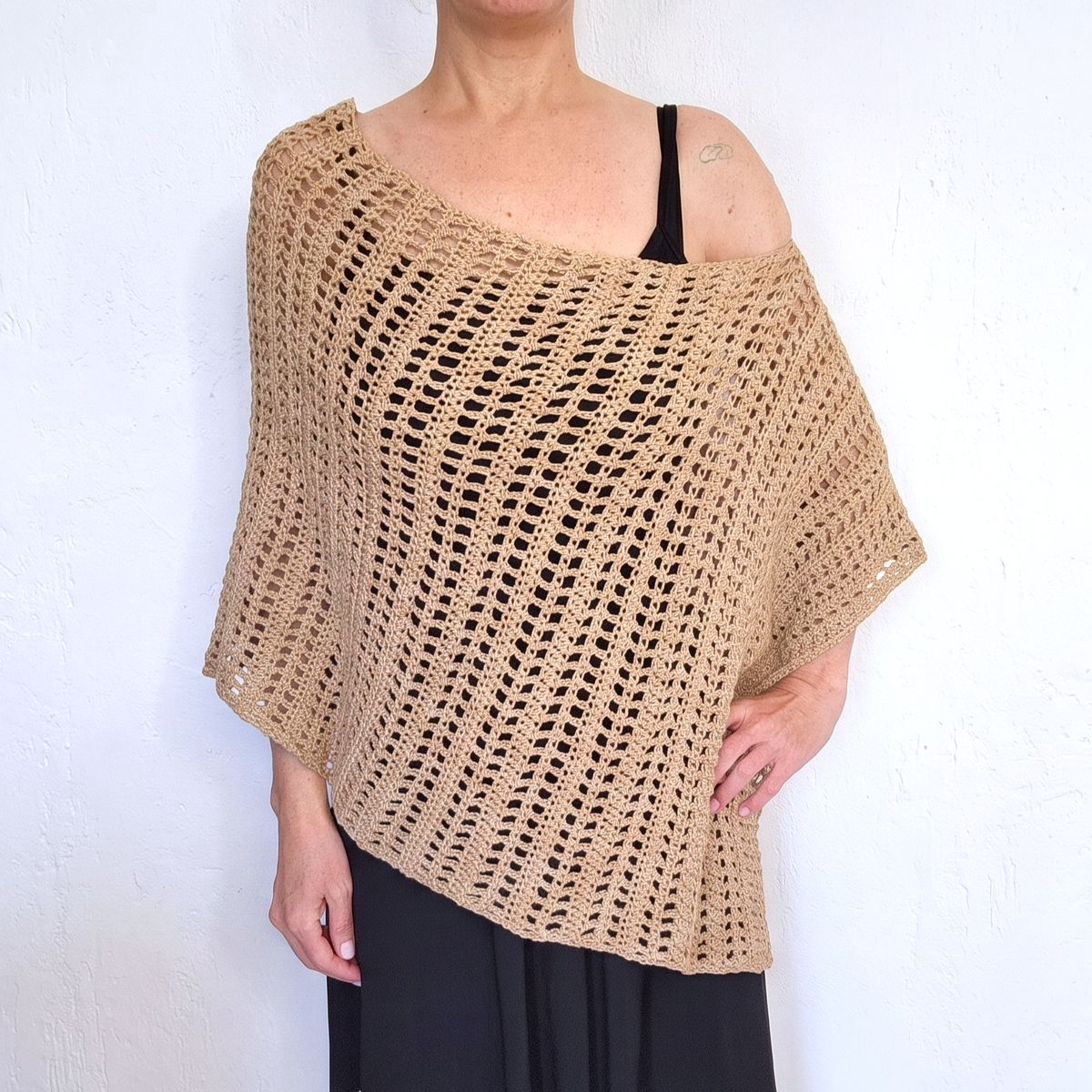 A woman wearing a beige open-knit crochet poncho over a black spaghetti strap top and black skirt, standing against a white textured wall.