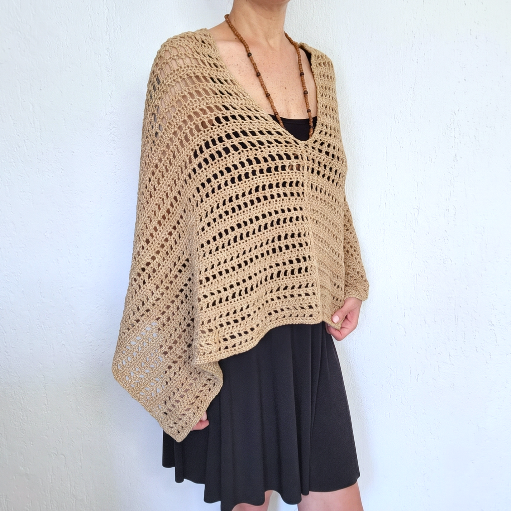A woman wearing a beige open-knit crochet poncho over a black top and black skirt, standing against a white textured wall.