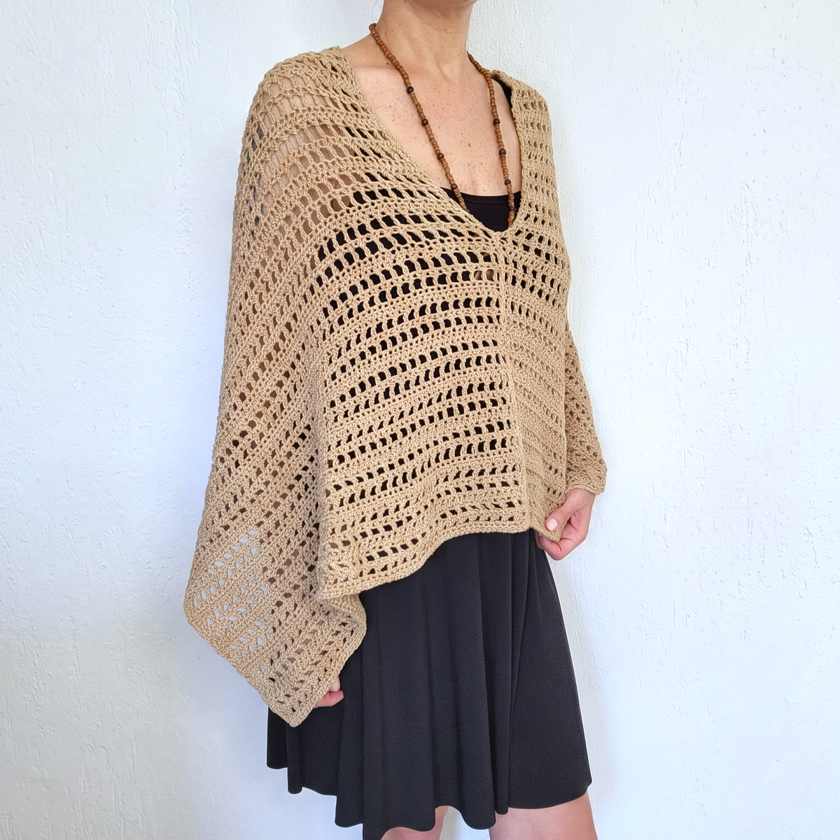 A woman wearing a beige open-knit crochet poncho over a black top and black skirt, standing against a white textured wall.