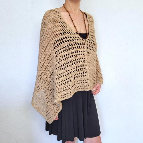 A woman wearing a beige open-knit crochet poncho over a black top and black skirt, standing against a white textured wall.