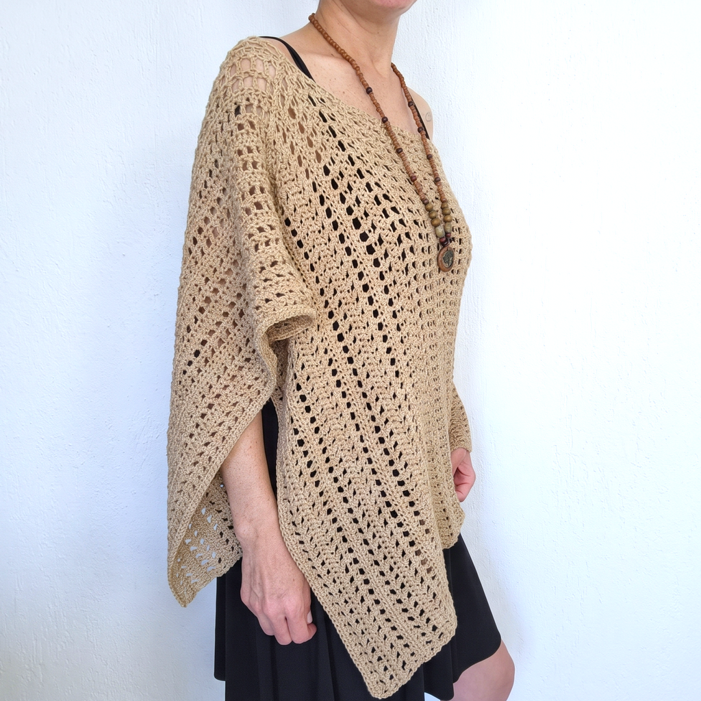 A woman wearing a beige open-knit crochet poncho-style top over a black dress, standing against a plain white wall.