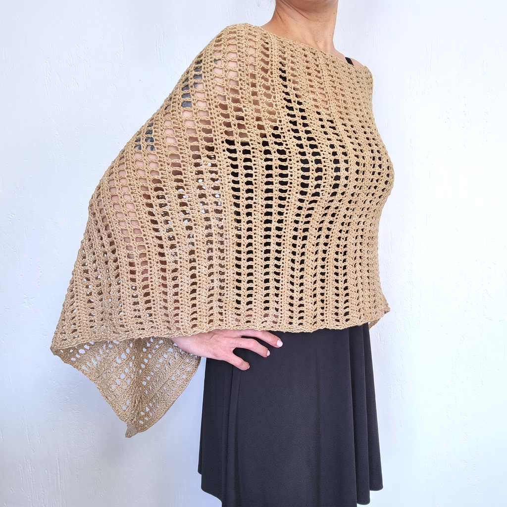 A beige open-knit crocheted poncho or shawl draped over a model's shoulders, paired with a black skirt, shown against a plain white wall.