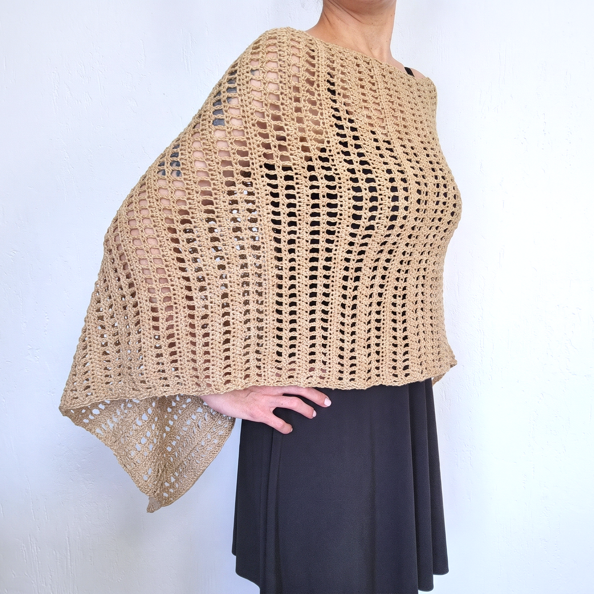 A beige open-knit crocheted poncho or shawl draped over a model's shoulders, paired with a black skirt, shown against a plain white wall.