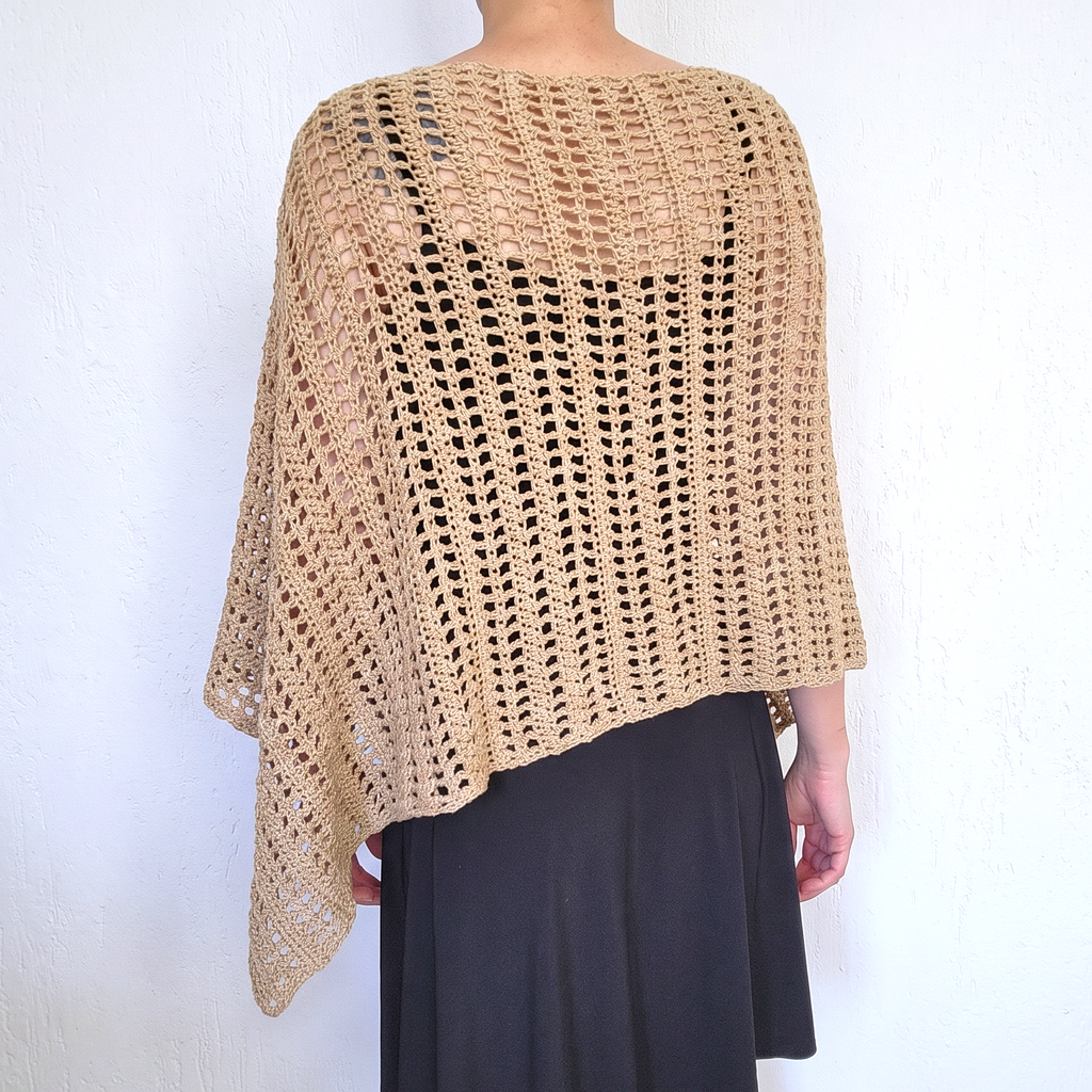 A person shown from the back wearing a loose, open-knit crocheted poncho in a warm beige/tan color over a dark skirt, standing against a plain white wall.